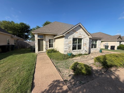 A house with a brown roof and a stone facade is surrounded by a well-maintained lawn and a sidewalk.