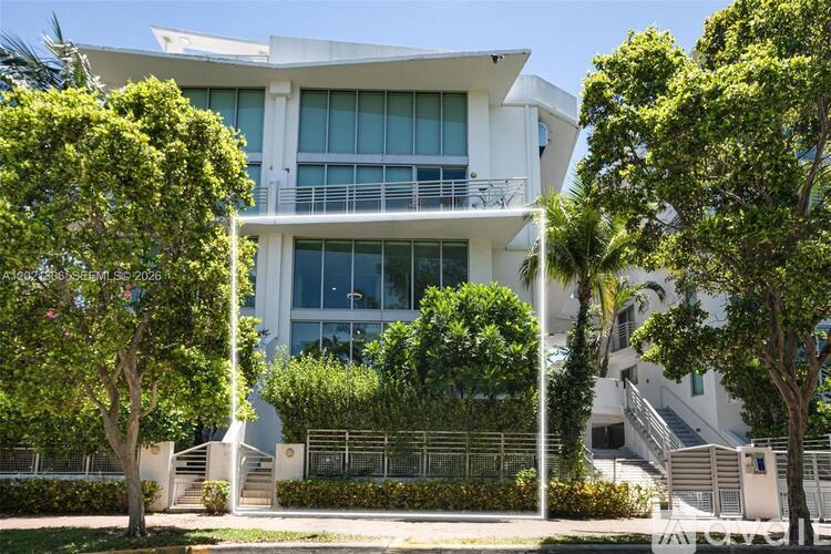 A modern white building with a balcony is surrounded by green trees.