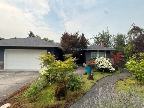 A house with a grey roof and a white garage door is surrounded by a garden with a gravel path.