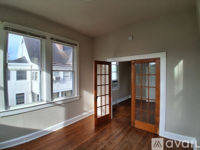 A room with a wooden floor, a window with a view of a building outside, and a door leading to another room.