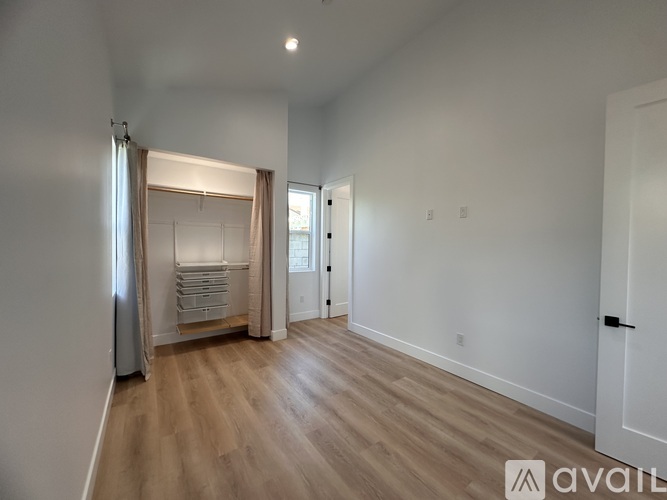 A room with wooden flooring and white walls, with a door on the right and a window with curtains on the left.