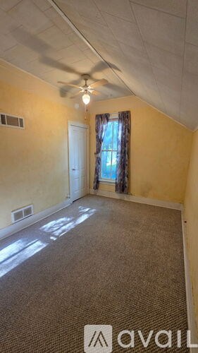 A room with a carpeted floor, a ceiling fan, and a window with curtains.