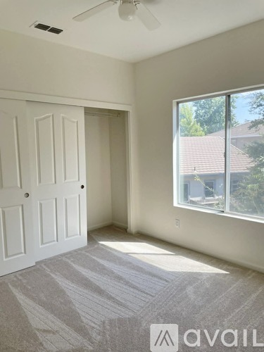 A room with a carpeted floor, a ceiling fan, and a large window with a view of a house and trees outside.