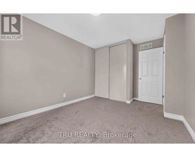 A bedroom with beige walls and carpeting, a white door, and a brokerage sign.