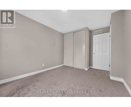 A bedroom with beige walls and carpeting, a white door, and a brokerage sign.