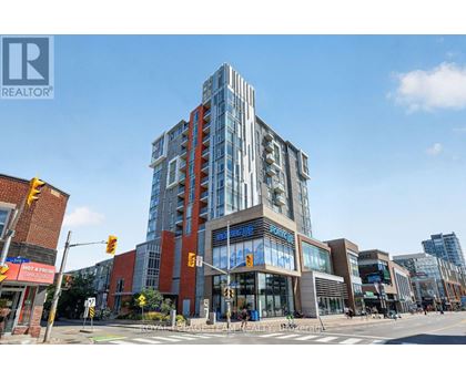 A modern multi-story building with glass windows and a sign that reads "REALTOR" is situated at an intersection with traffic lights and pedestrian crossings.