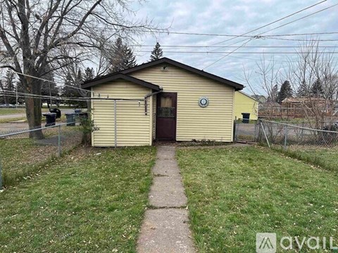 A small yellow building with a brown door is surrounded by a grassy area and a fence.