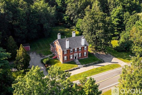 A large red brick house with a black roof and chimneys is surrounded by green trees.