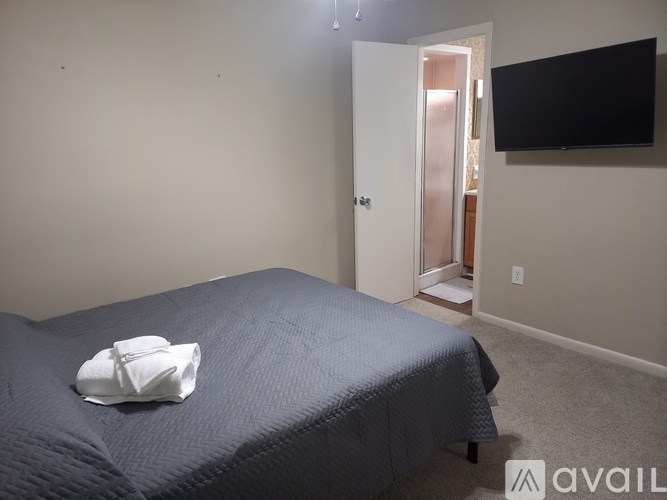 A bed with a blue comforter and a white pillow is in a room with a television and a doorway leading to another room.