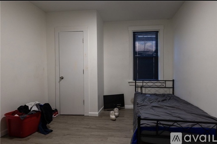 A bedroom with a bed, a window with blinds, a door, a red storage box, and a pair of shoes.