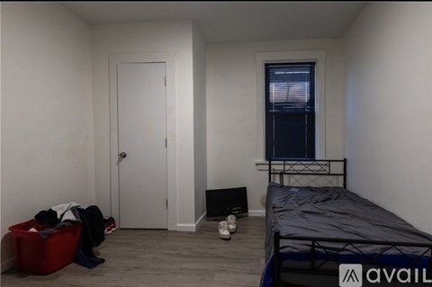 A bedroom with a bed, a window with blinds, a door, a red storage box, and a pair of shoes.