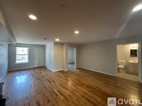 A room with wooden flooring and white walls, with a bathroom visible in the background.