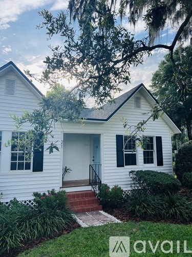 A white house with a black door and windows is surrounded by greenery.