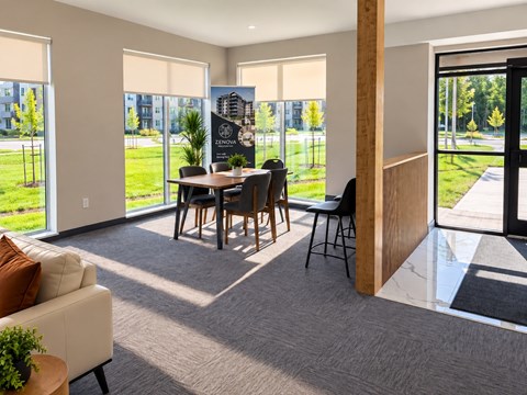 A modern living room with a dining table and chairs, a sofa, and a view of a green outdoor area through large windows.