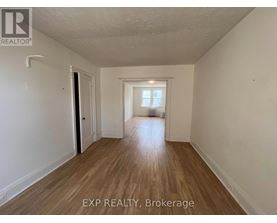 A room with wooden flooring and white walls, with a door on the left and a window on the right.