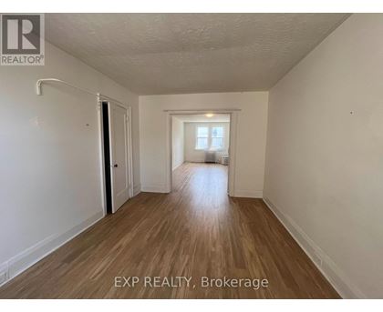 A room with wooden flooring and white walls, with a door on the left and a window on the right.