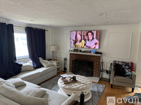 A living room with a grey couch, a white ottoman, a fireplace, and a television on the wall.