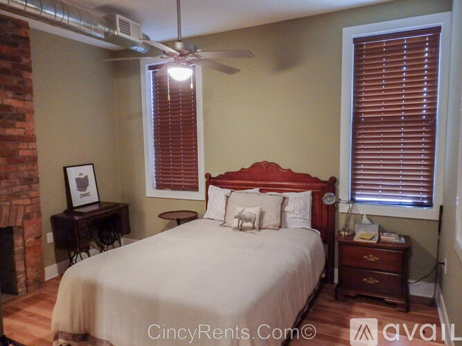 A bedroom with a red headboard bed, a small table with a lamp and a picture frame on it, and a window with blinds.