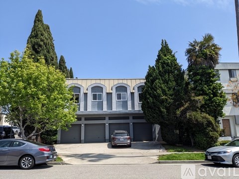 A modern house with a grey garage door is surrounded by trees and parked cars.