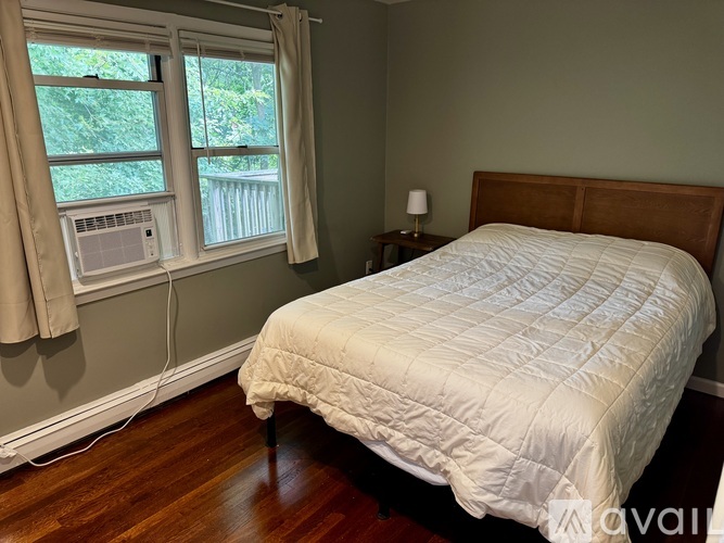 A bedroom with a bed, a window with a fan, and a nightstand with a lamp.