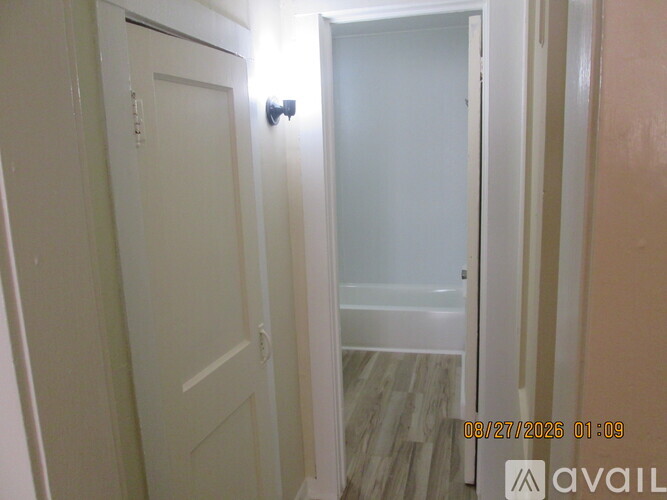 A room with a brown floor and white walls has a doorway leading to another room.