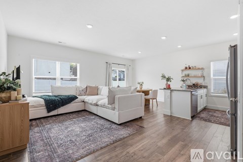 A well-lit, spacious living room with a white sofa set, wooden floors, and a kitchen area in the background.