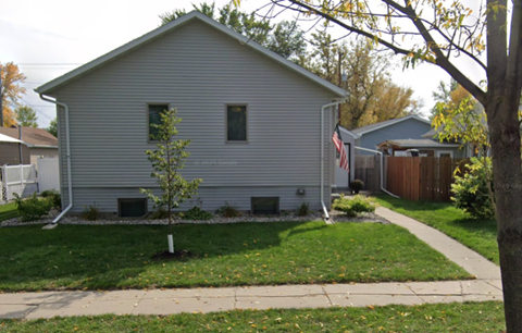 A small house with a grey siding and a small tree in front of it.