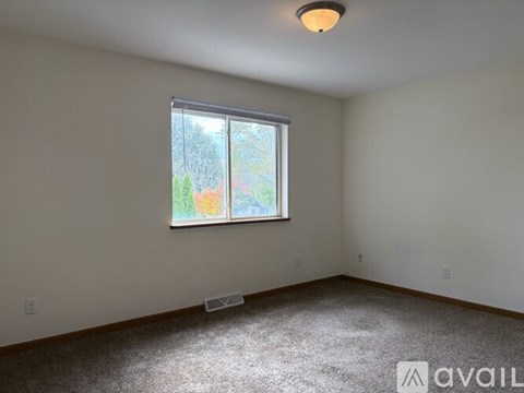 A room with a carpeted floor, a window with a view of trees and a house, and a ceiling light fixture.