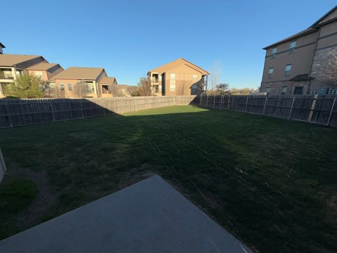 A backyard with a concrete pathway leading to a fenced area with a grassy field and a building in the background.