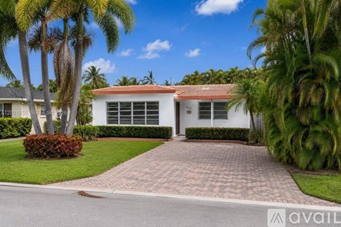 A house with a red roof and white walls is surrounded by greenery and palm trees.