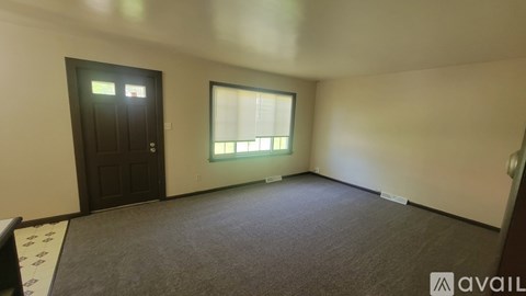 A room with a carpeted floor, a window with blinds, and a door with a sign above it.