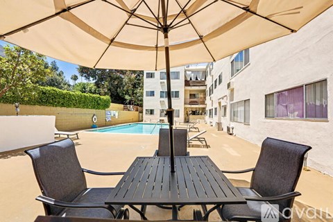 A table with chairs and an umbrella is in the foreground of a pool area.