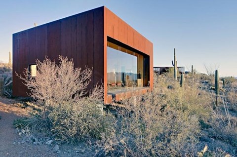 A modern house with a large window and a brown exterior is surrounded by desert vegetation.