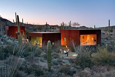 A modern house with a red exterior is surrounded by cacti and other desert vegetation.