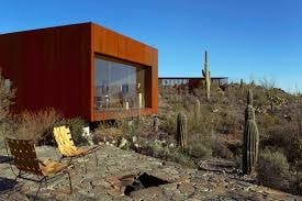 A modern house with a red exterior is surrounded by cacti and chairs are placed outside.