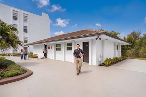 A man in a black shirt and khaki pants is walking in front of a white building with a brown roof.