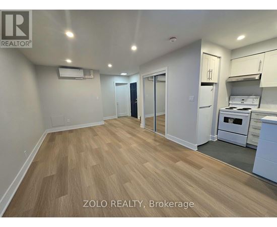 A well-lit, empty room with wooden flooring and white walls, featuring a kitchenette with white appliances.