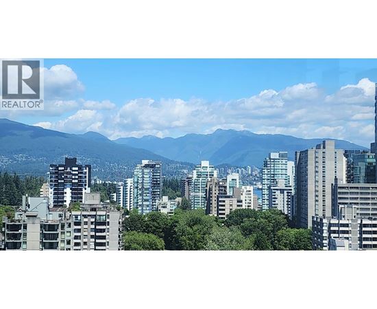 A cityscape with buildings and trees in the foreground and mountains in the background.