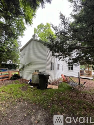 A white house with a chimney is surrounded by trees and has a trash can in front of it.