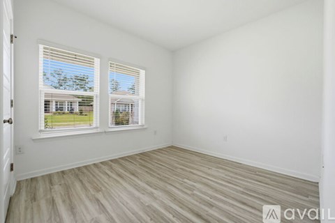A room with white walls and wooden flooring, with two windows showing a view of a house and trees outside.