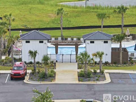 A red car is parked in a parking lot with a pool and two white buildings in the background.