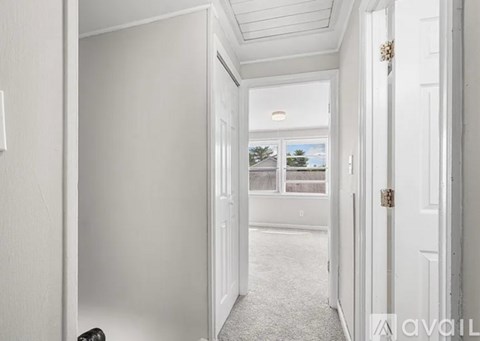 A hallway with white walls and a carpeted floor leading to a window with a view of trees and a house.