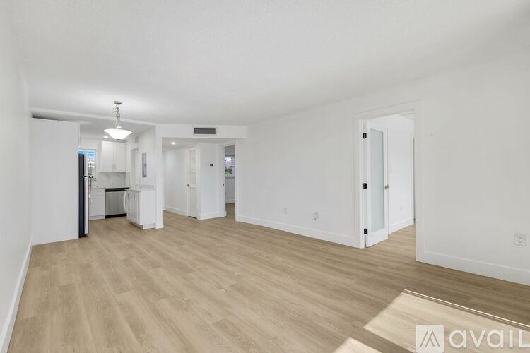 A spacious room with wooden flooring and white walls, with a kitchen area visible in the background.