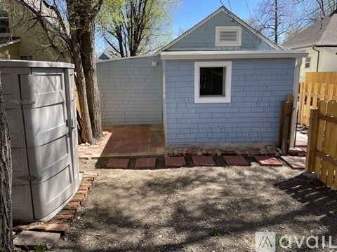 A small blue building with a white window is surrounded by a brick pathway and a wooden fence.