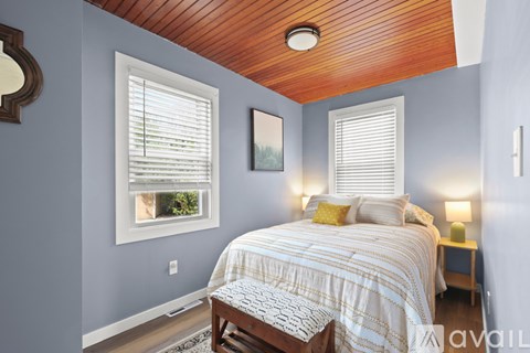 A bedroom with a bed, a window with blinds, a nightstand with a lamp, and a picture on the wall.
