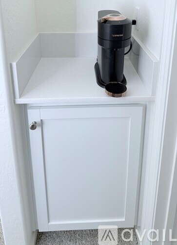 A black coffee maker is on a white shelf inside a white cabinet.