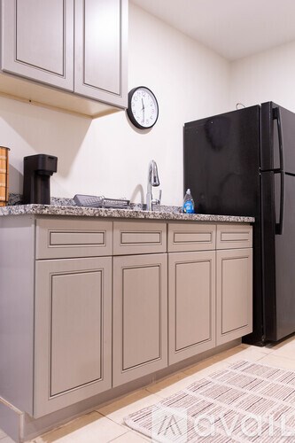 A black refrigerator sits next to a counter with a clock on the wall.