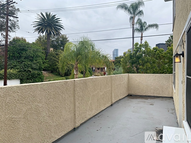 A balcony with a concrete floor and a wall on one side, surrounded by greenery and buildings in the distance.