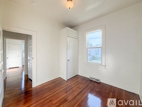 A room with wooden floors and white walls, with a window showing a view of buildings outside.
