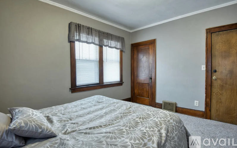A bedroom with a bed, a window with blinds, a wooden door, and a wall-mounted air conditioner unit.